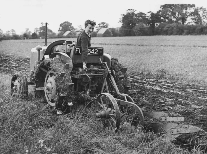 Black and white image from the WWII era of a woman in a field on a tractor.